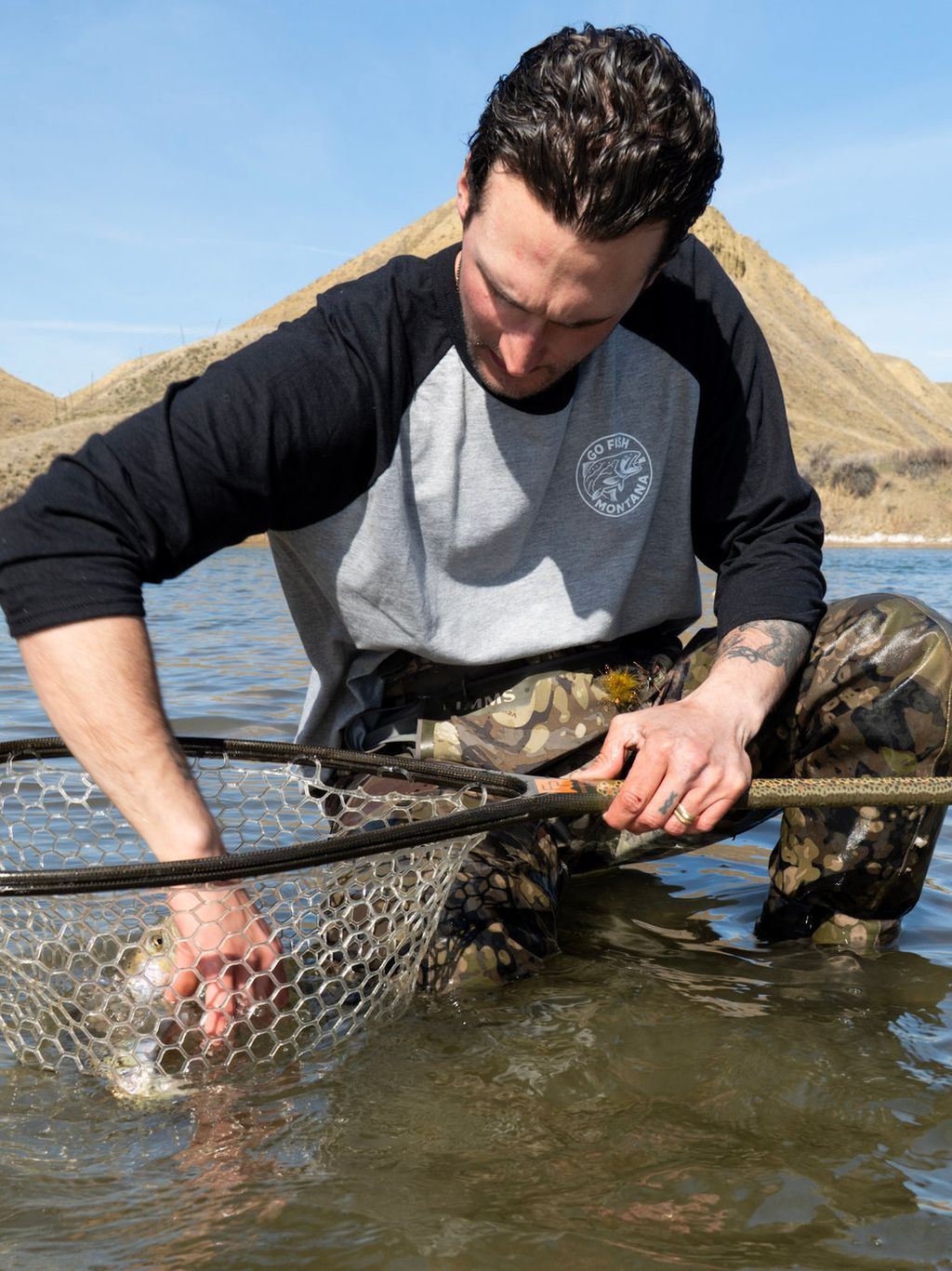 Angler in a GFM baseball tee leaning over a landing net in shallow river water