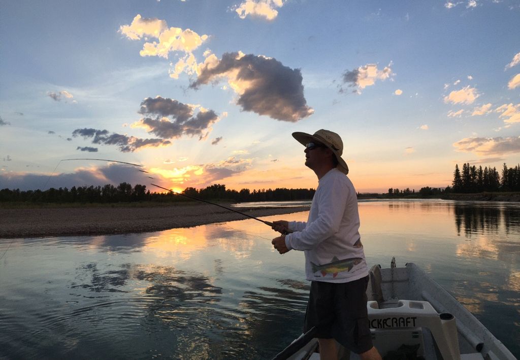 An angler stands in a boat with a fly rod as the sun sets over calm water